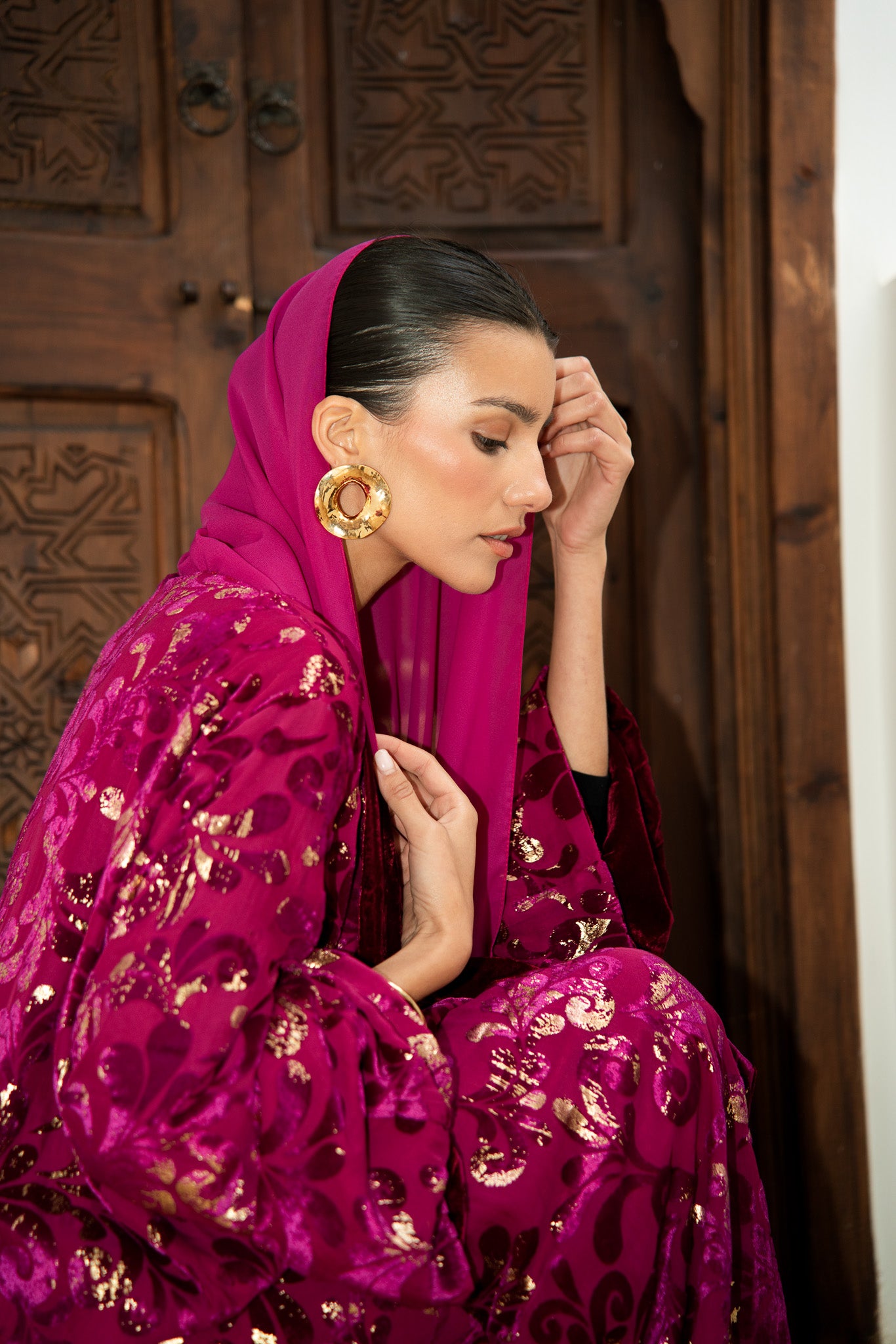 Maroon velvet open abaya with gold floral detail throughout, worn with matching maroon premium chiffon hijab, showing elegant flowing silhouette against ornate wooden door backdrop